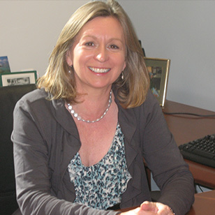 Dr. Gina Piane sits at a desk. She is wearing a brown sweater and has medium length blonde hair.