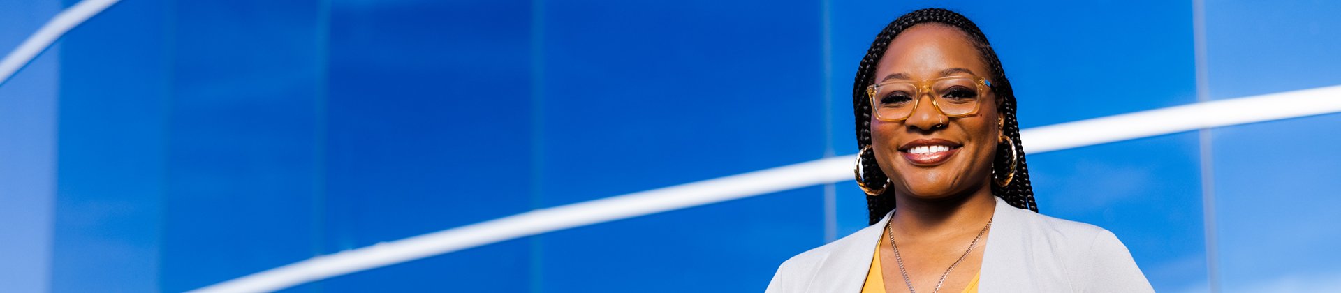 Britnnei P. wears clear glasses and hoop earrings, she is standing in front of a blue background