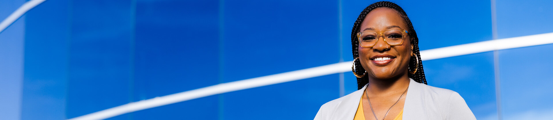 Brittnei P., a woman who wears glasses and small braids, smiles for the camera in front of a blue background.