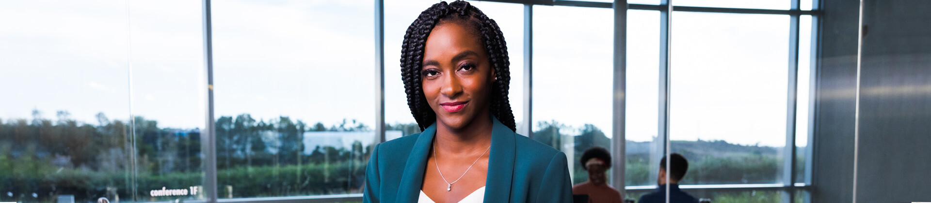 Spshelle R. stands in front of a glass window. She is wearing a teal blazer and has long braids.