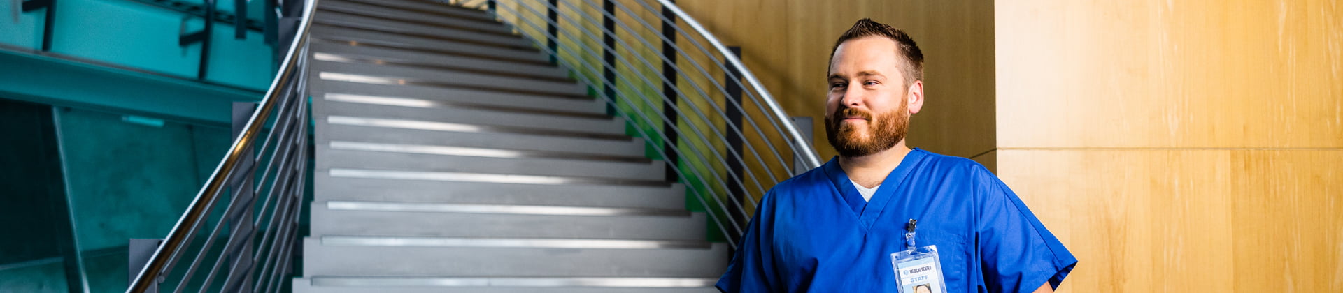 Kyle W., Class of 2018 stands at the base of a staircase. He is wearing blue scrubs and has short hair with a reddish beard.