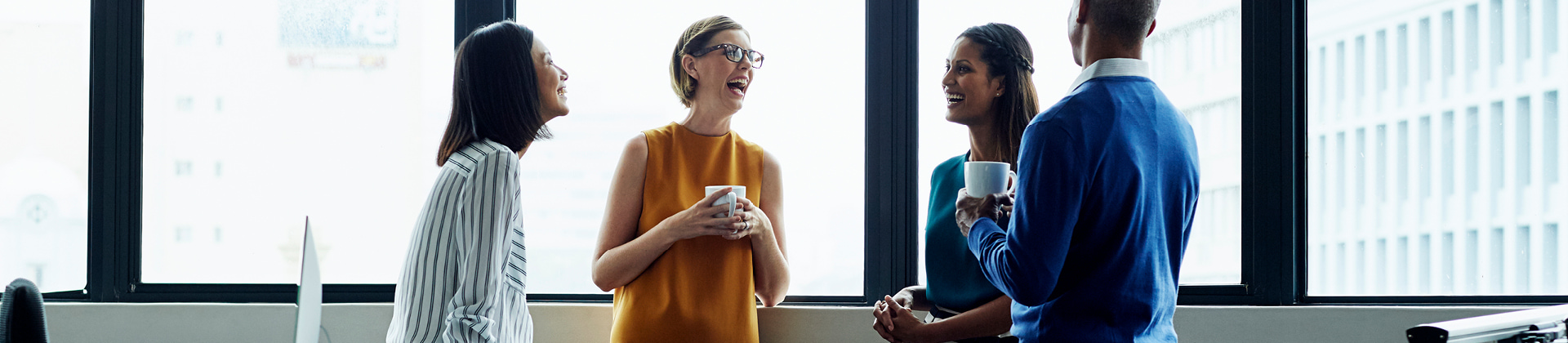 four individuals in a semicircle engaged in conversation, two of them with coffee mugs, three women are smiling, one man has back turned to camera