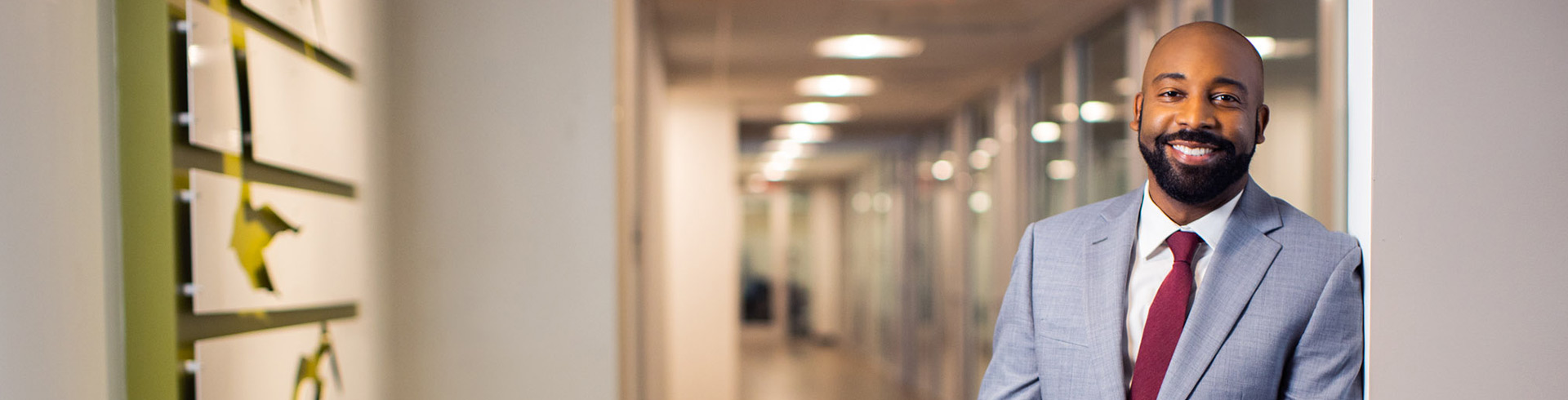 man with a beard in a gray suit smiles in a hallway